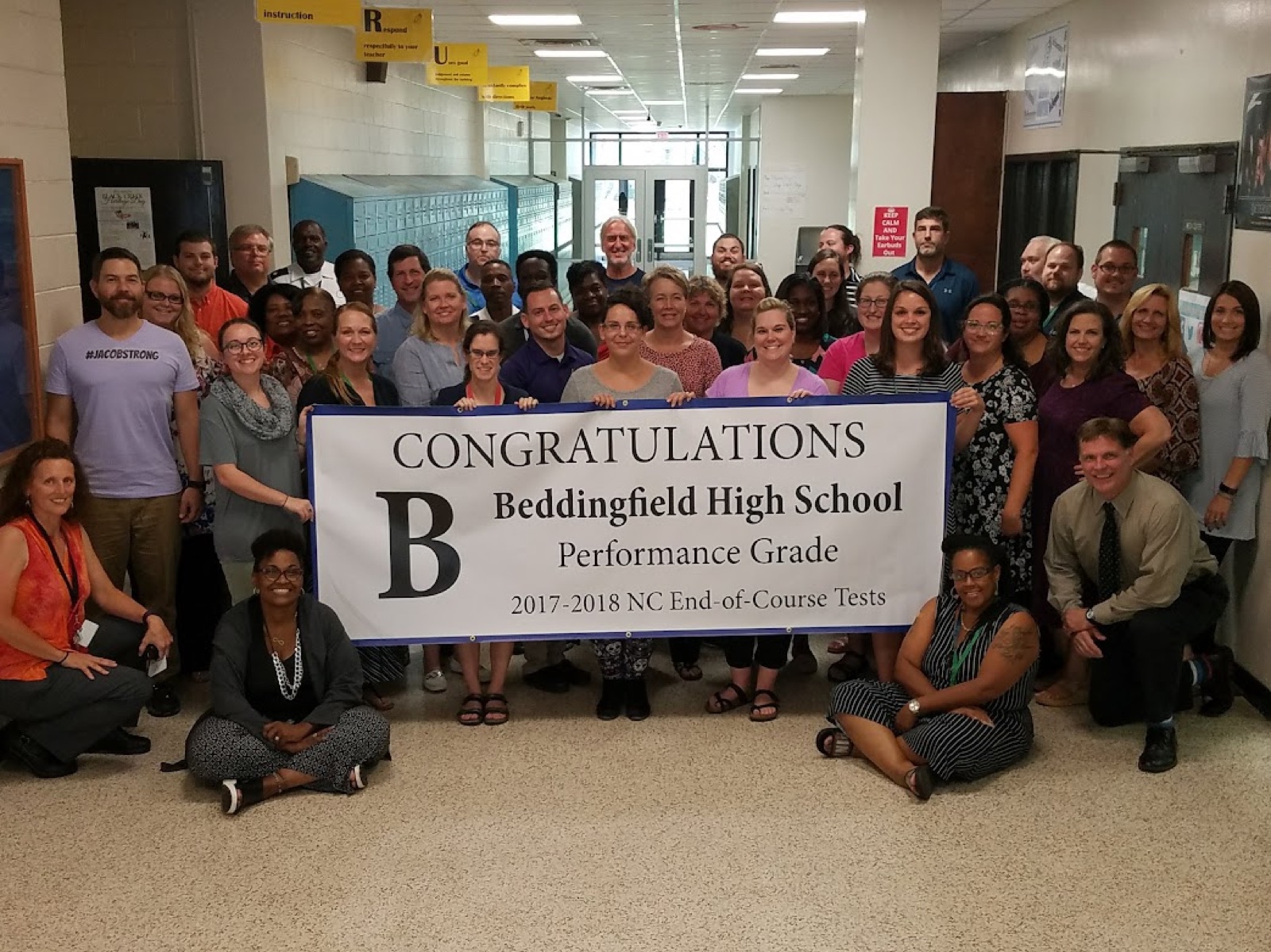 Roughly fifty Beddingfield High School staff members gathered in a hallway around a banner reading Congratulations B, Beddingfield High School Performance Grade, 2017-2018 NC End-of-Course Tests.