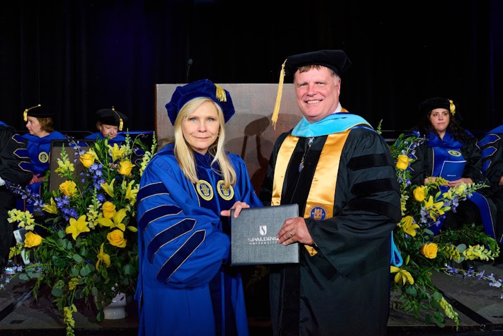 F.T. Franks in black doctoral regalia and tam receives his diploma folder on a commencement stage at Spalding University.