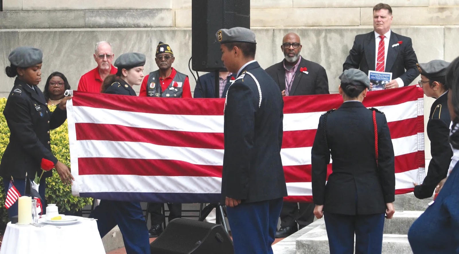 JROTC cadets hold a large American flag at an outdoor ceremony while F.T. Franks speaks from a podium beside a group of veterans.