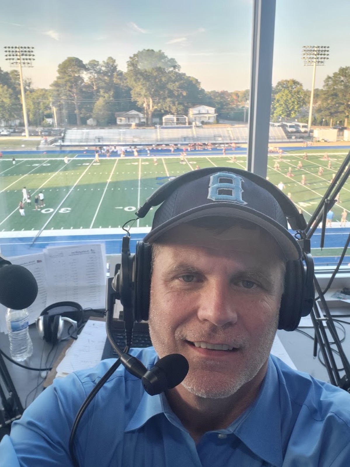 F.T. Franks in a broadcast headset and a Beddingfield B cap in a press box, with a football field and players warming up in the evening light behind him.