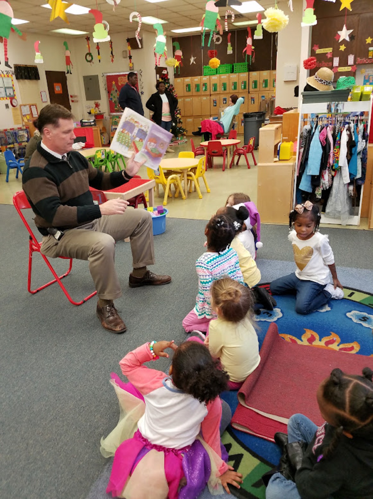 F.T. Franks sits on a small red chair in a decorated preschool classroom, holding up a picture book for a group of children seated on the rug.